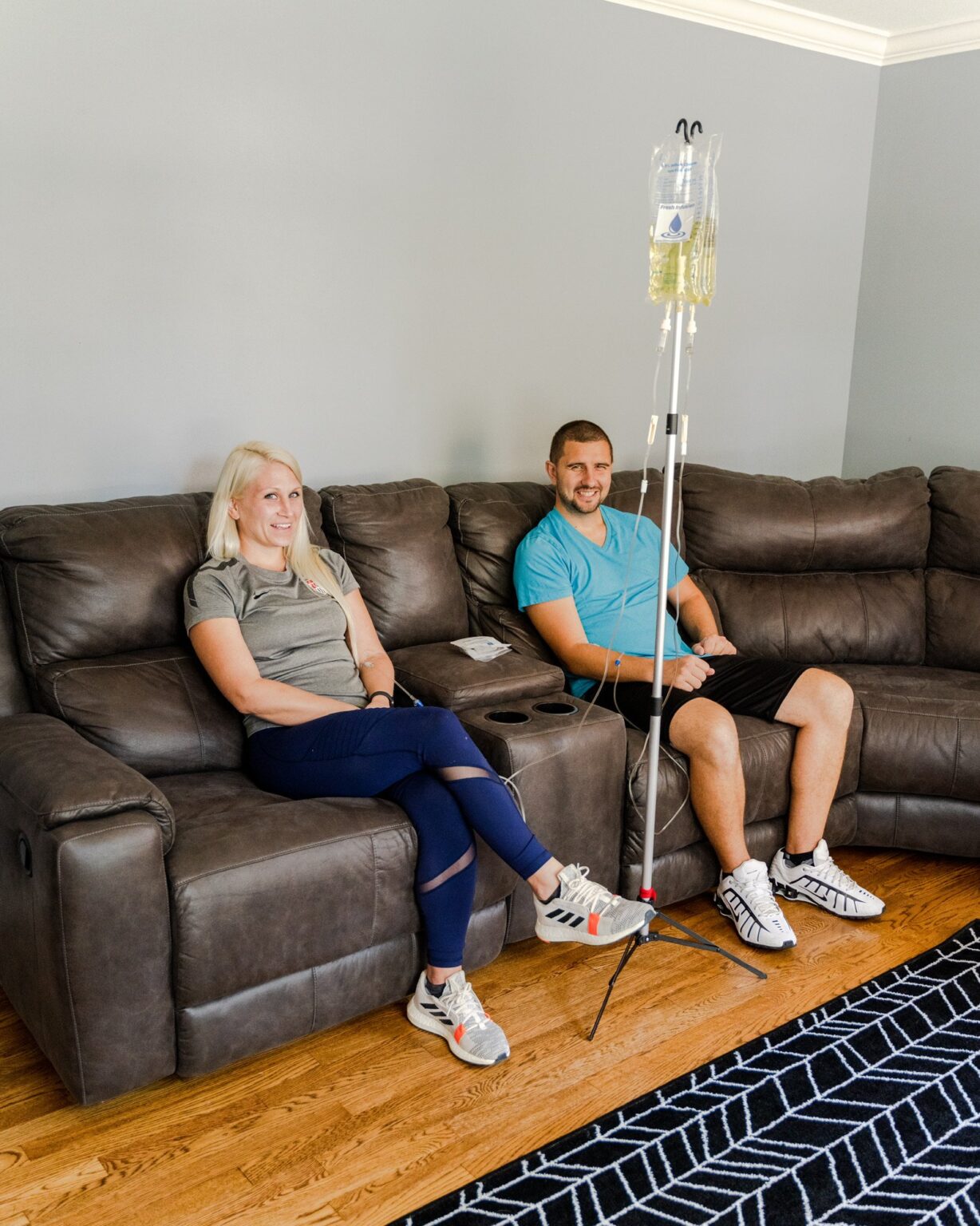 Two adults receiving IV therapy at home on a sofa, home medical care supplies visible.