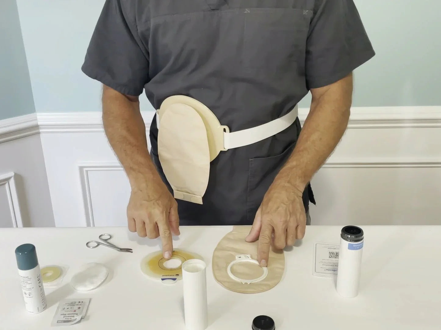 Person demonstrating two-piece ostomy pouch system with medical supplies on table.