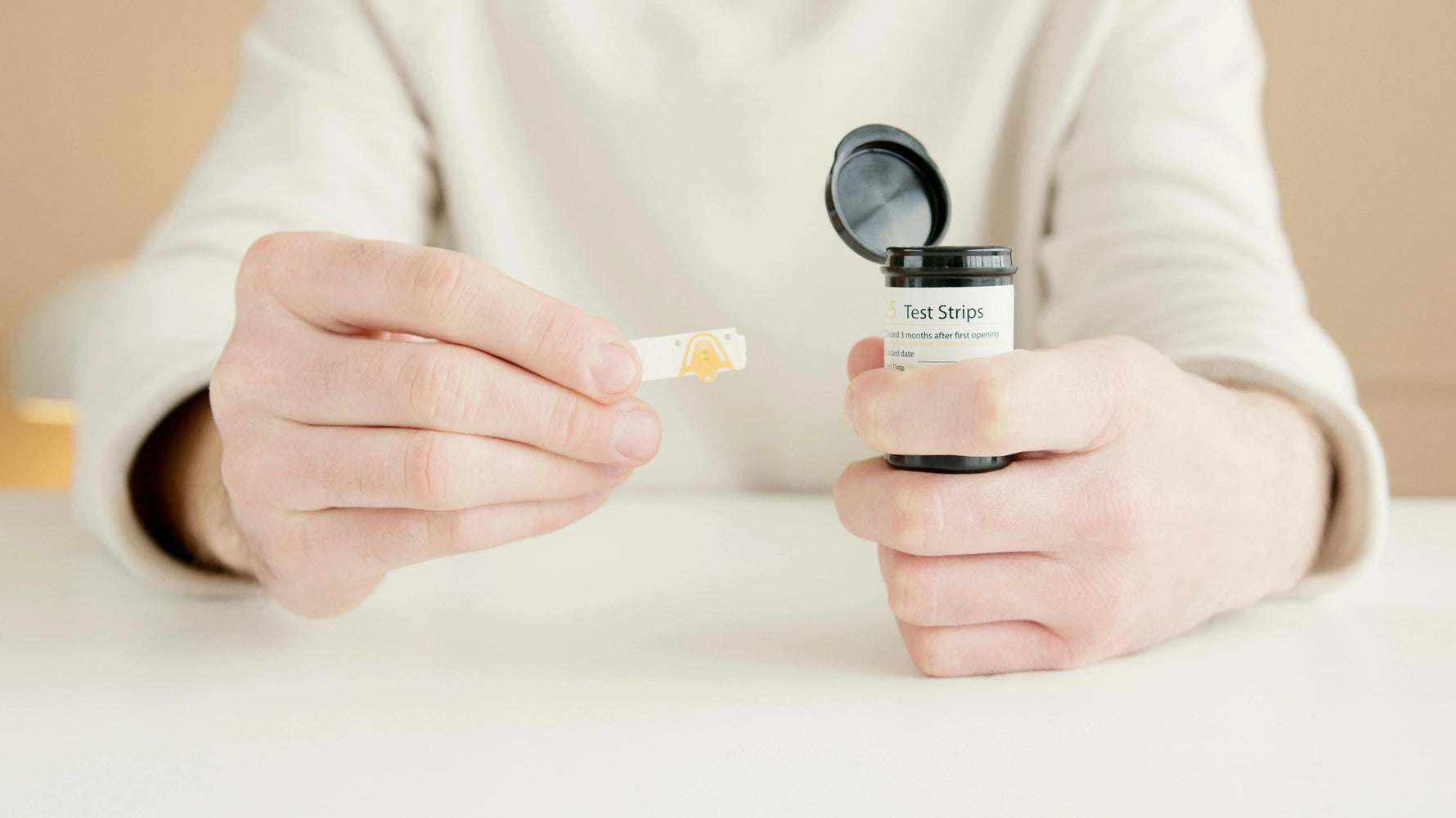 Person holding medical test strips container and a single test strip on a white table