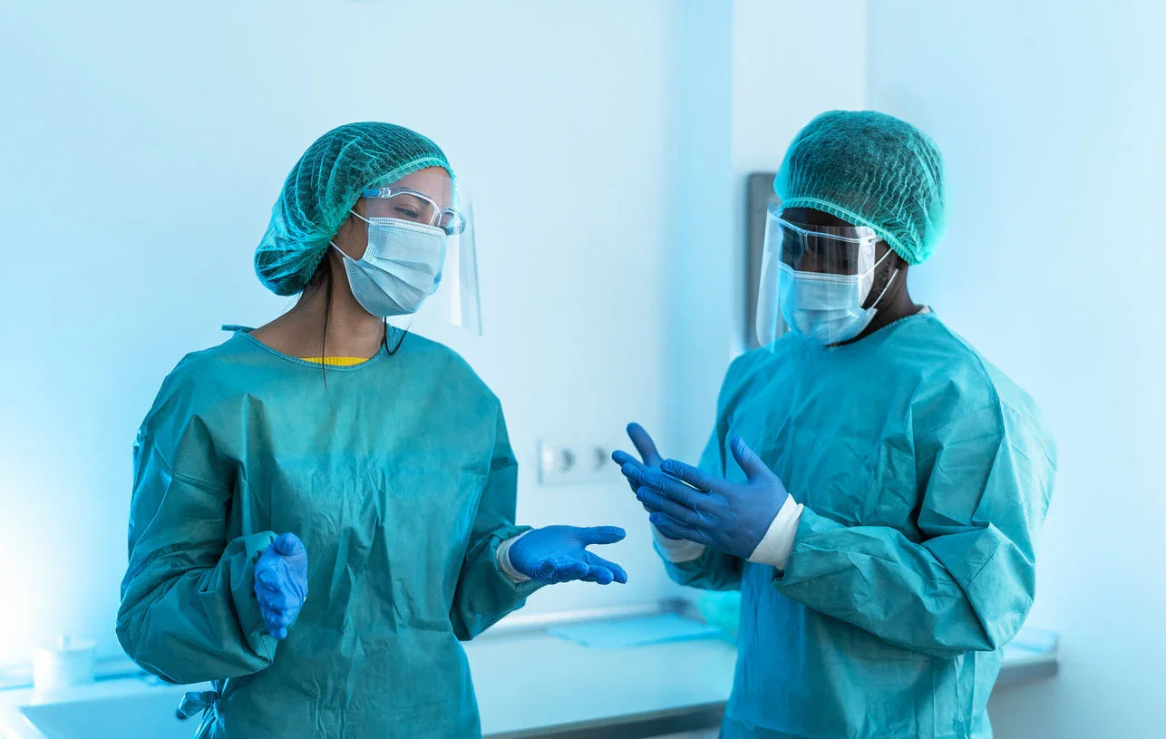 Healthcare workers in PPE gowns, gloves, masks, and face shields talking in a medical setting.