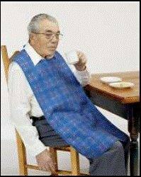 Elderly man wearing an adult bib seated at table, drinking, with home care supplies visible.